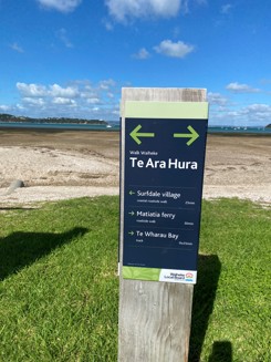 A sign with two arrows pointing in opposite directions with three locations: Surfdale distance, Matiatia ferry and Te Wharau Bay with the walking distance to those destinations.