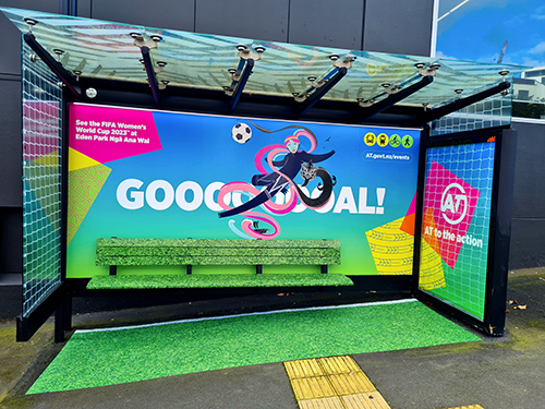 Image of bus shelter on Victoria Street in Auckland city, that has been re-purposed as a football goal including our Auckland Transport wāhine mahi toi (artwork), fake grass and goal netting.