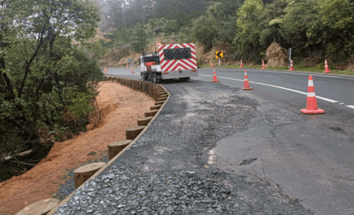 An asphalt truck works on a road marked with orange cones. A retaining wall made of wood piles reinforces the side where asphalt is being laid