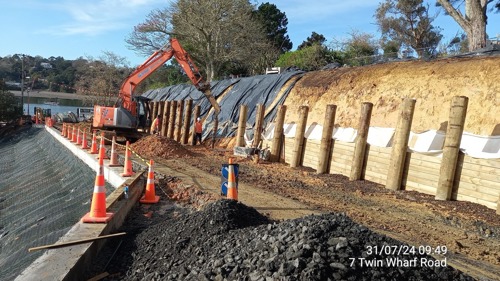 Construction crew and machinery installing timber poles on a retaining wall alongside a dirt bank. A lower bank is covered with mesh and marked off with orange cones, and a pile of gravel is in the foreground.