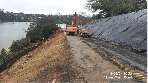 Digger working on mud and concrete road with black plastic sheeting covering nearby sloping bank