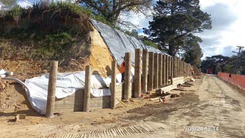 A timber retaining wall with all piles now in. A construction worker stands behind the piles, installing the timber lagging. Plastic sheeting stops rainwater from further eroding the bank.