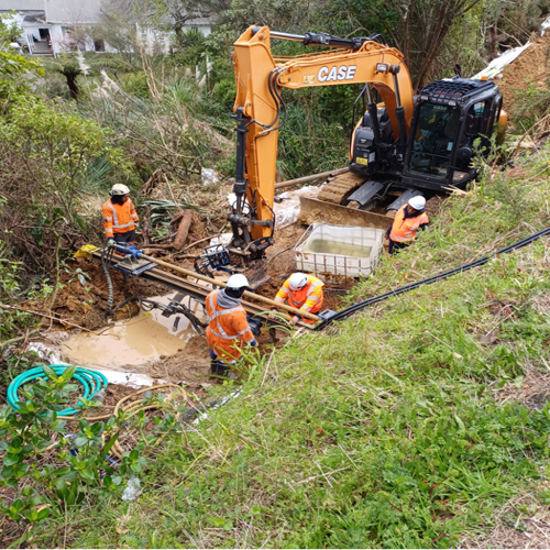 Drainage work is underway. An excavator is supporting drilling equipment being used by 4 work personnel in wet and muddy conditions.