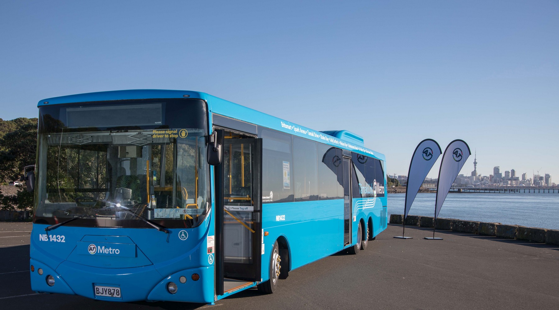 Tāmaki Link blue bus parked on the side of the road with the Auckland skyline in the background.