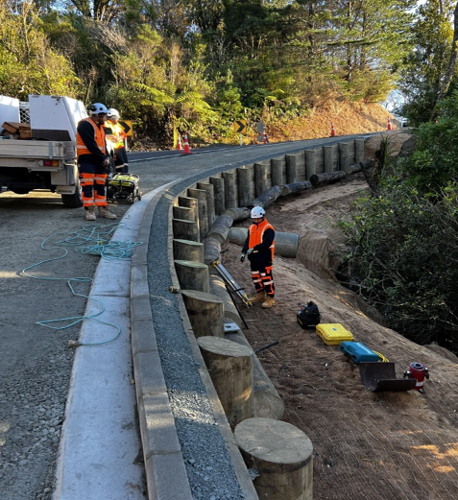 2 road workers stand on the road with a generator and equipment while a third stands below the road at the bottom of the new timber retaining wall, with equipment used for the finishing touches of work.