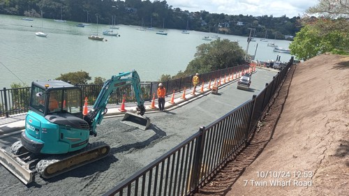 An excavator spreads the fine metal layer on the road area. A roller operates in the background to compact the metal prior to final asphalt work. To the right of the image, the bank area has fresh topsoil, ready for planting.